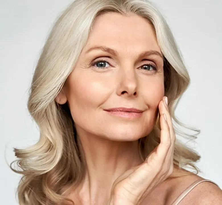 senior woman with light blonde curly hair posing with her hand on her cheek in front of a grey background