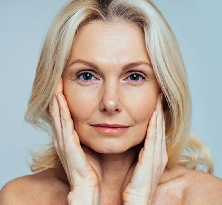 Senior woman with blonde and grey hair her hands on her cheeks posing in front of a blue background