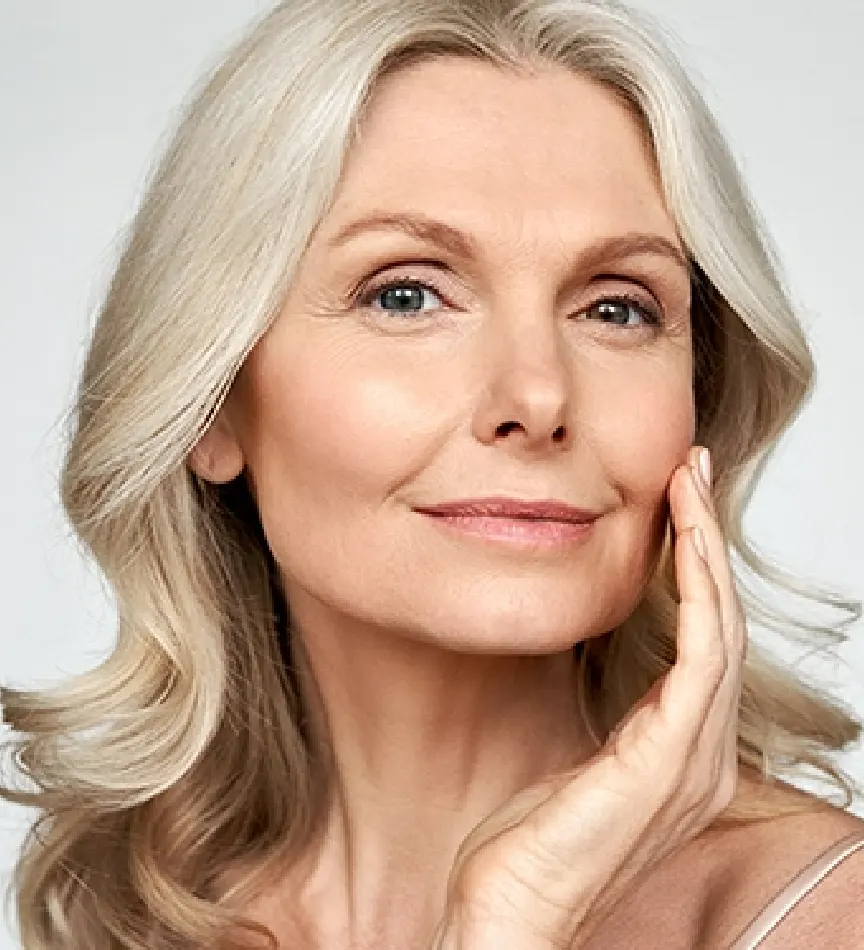 senior woman with light blonde curly hair posing with her hand on her cheek in front of a grey background