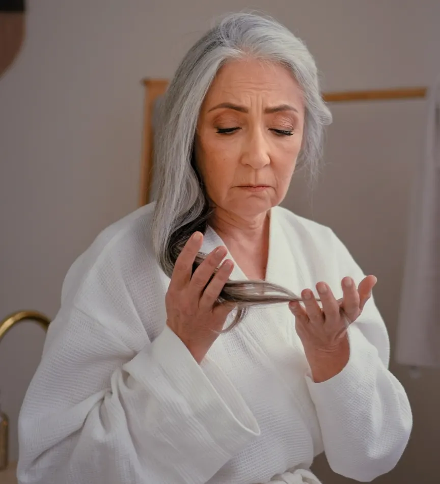 Senior Caucasian woman wearing a bathrobe, gently touching her gray, long, smooth hair, expressing concern about hair loss, thinning hairline, baldness, and the effects of aging, such as alopecia.