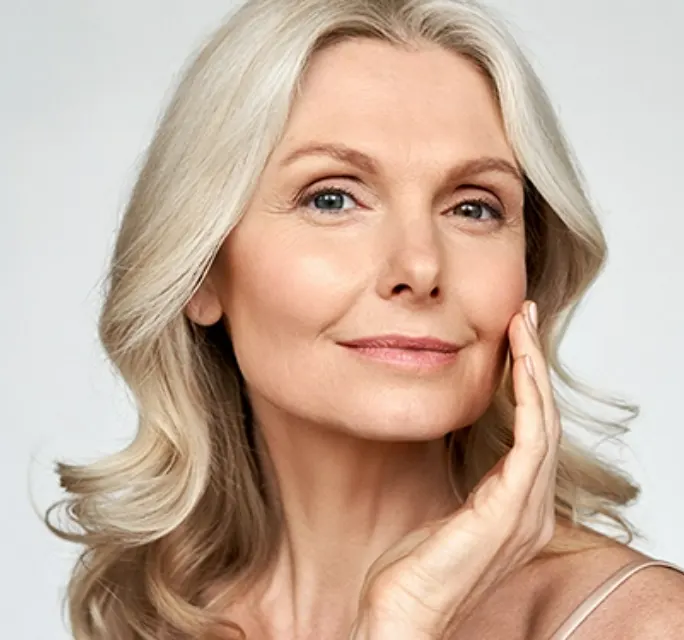 senior woman with light blonde curly hair posing with her hand on her cheek in front of a grey background