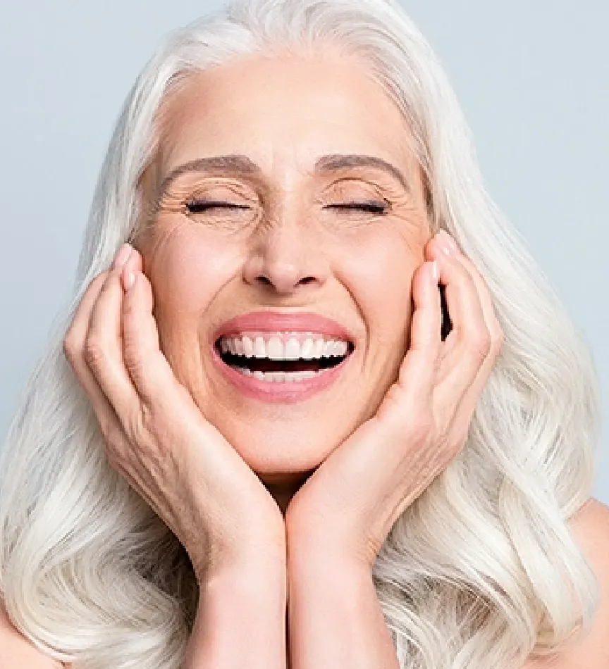 senior woman smiling with grey thick hair and her hands are placed on her cheeks as her eyes are closed in front of a grey background