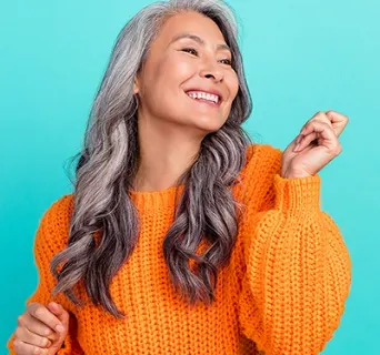 older woman with curly gray hair smiling and dancing wearing an orange sweater in front of a teal background