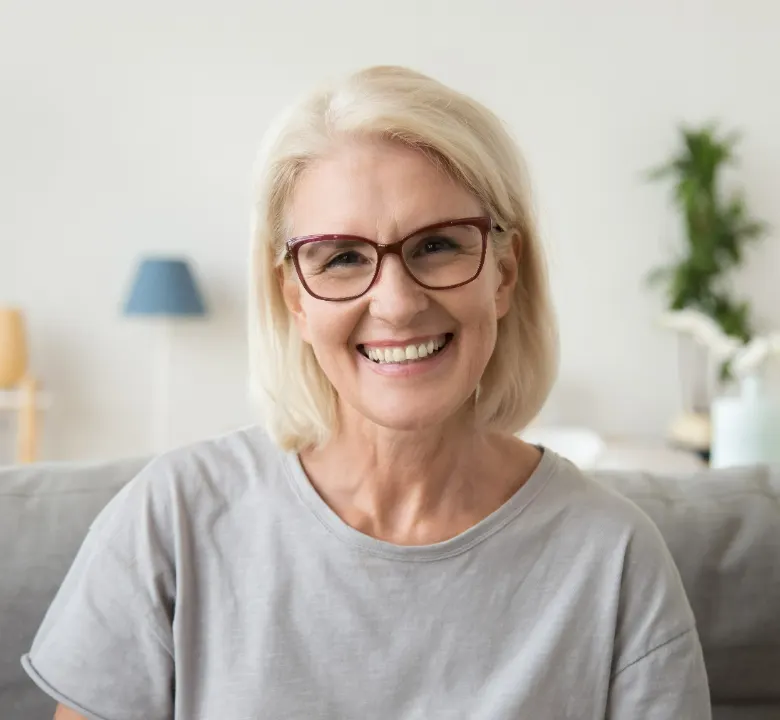 Smiling middle aged mature grey haired woman looking at camera while sitting on sofa in living room