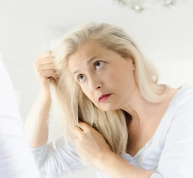 Upset senior woman looking through her thin hair as she stands in her bedroom looking in the mirror