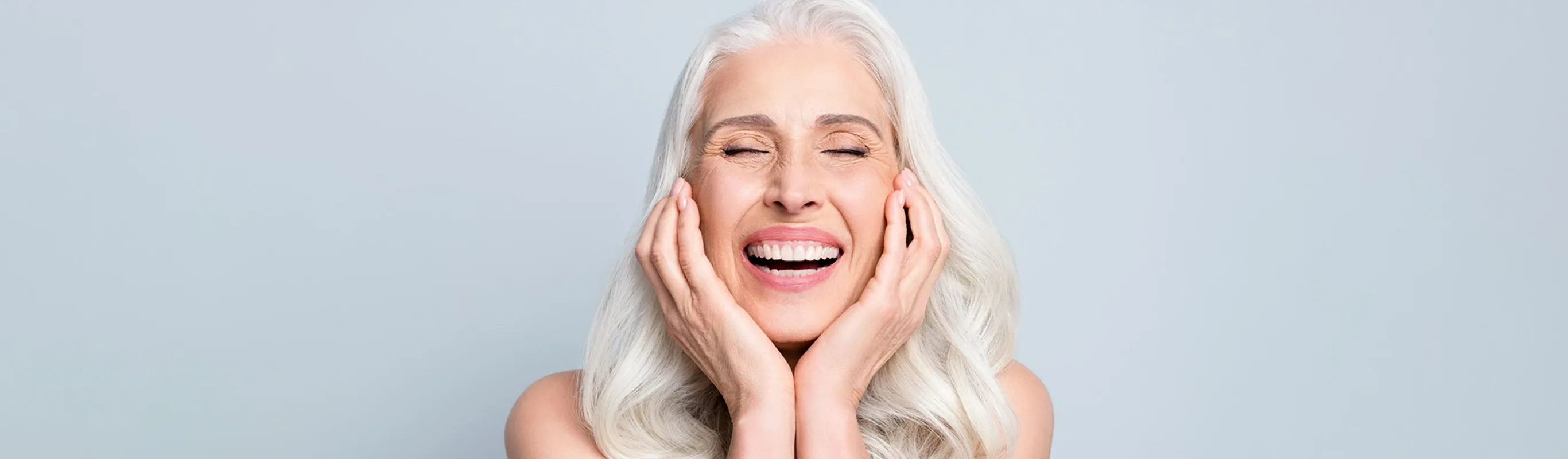 senior woman smiling with grey thick hair and her hands are placed on her cheeks as her eyes are closed in front of a grey background