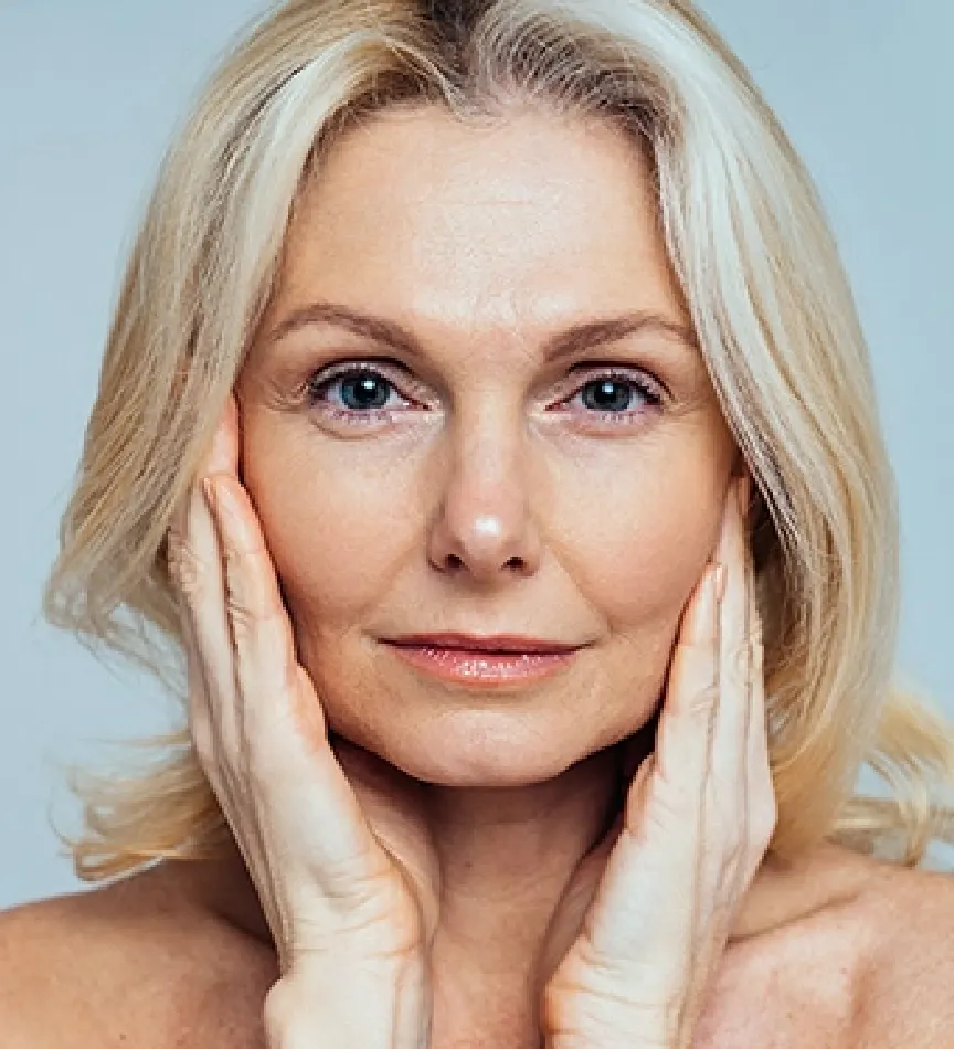 Senior woman with blonde and grey hair her hands on her cheeks posing in front of a blue background