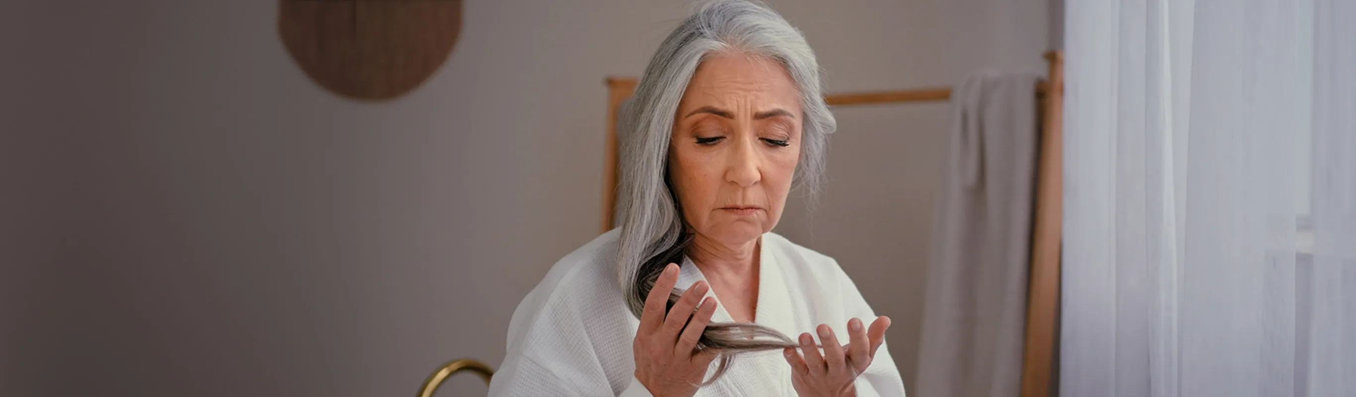 Senior Caucasian woman wearing a bathrobe, gently touching her gray, long, smooth hair, expressing concern about hair loss, thinning hairline, baldness, and the effects of aging, such as alopecia.