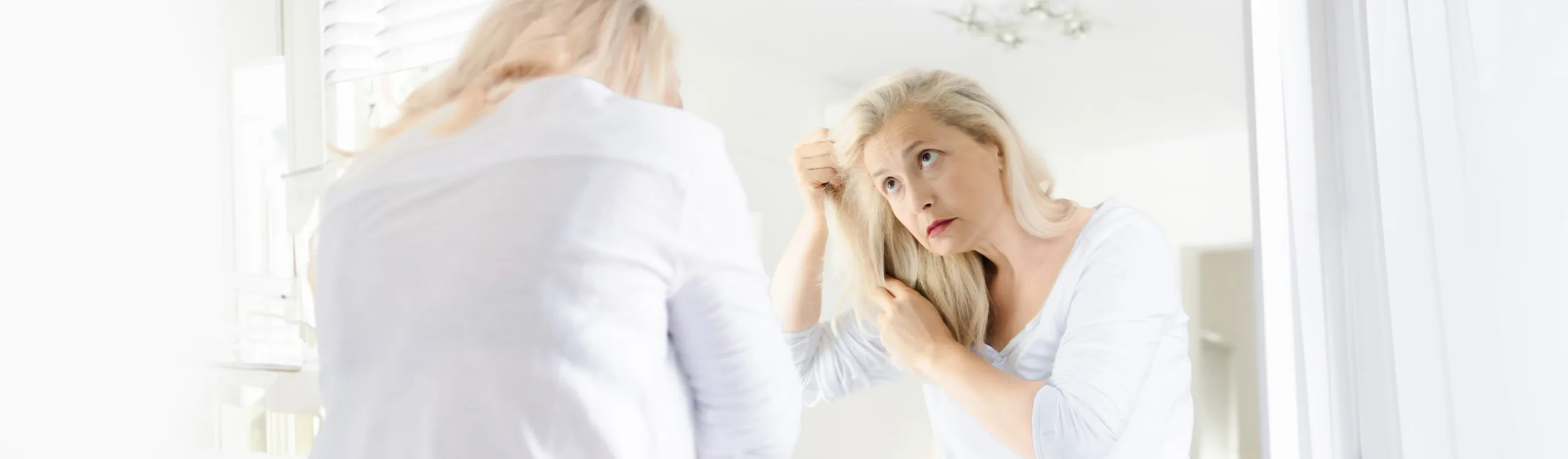 Upset senior woman looking through her thin hair as she stands in her bedroom looking in the mirror