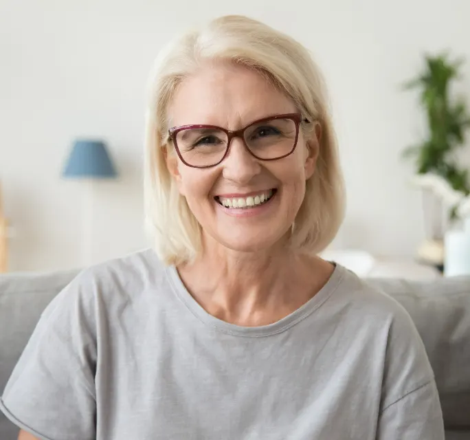 Smiling middle aged mature grey haired woman looking at camera while sitting on sofa in living room