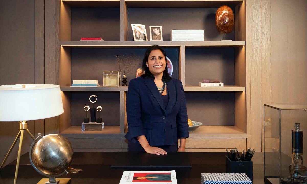 a person smiling in front of a desk with a lamp and bookshelf
