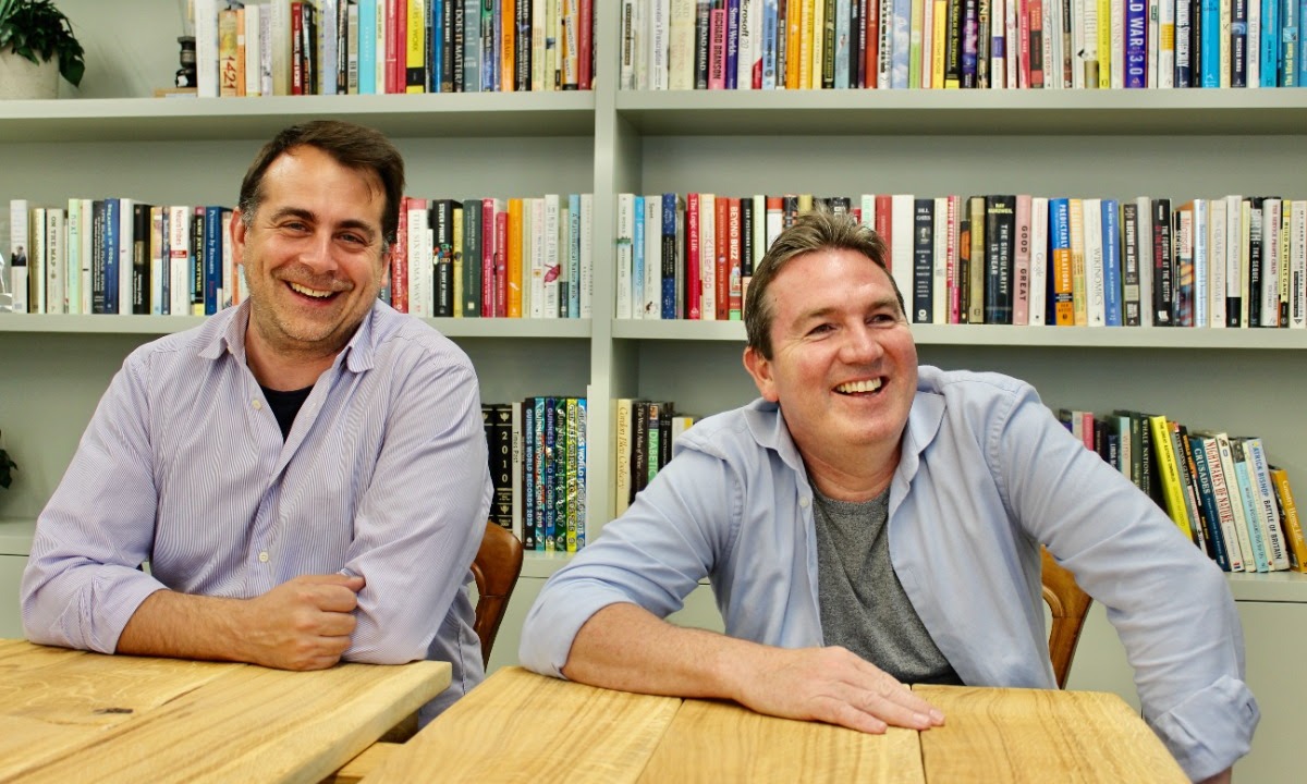 a couple of men sitting at a table in front of a bookshelf