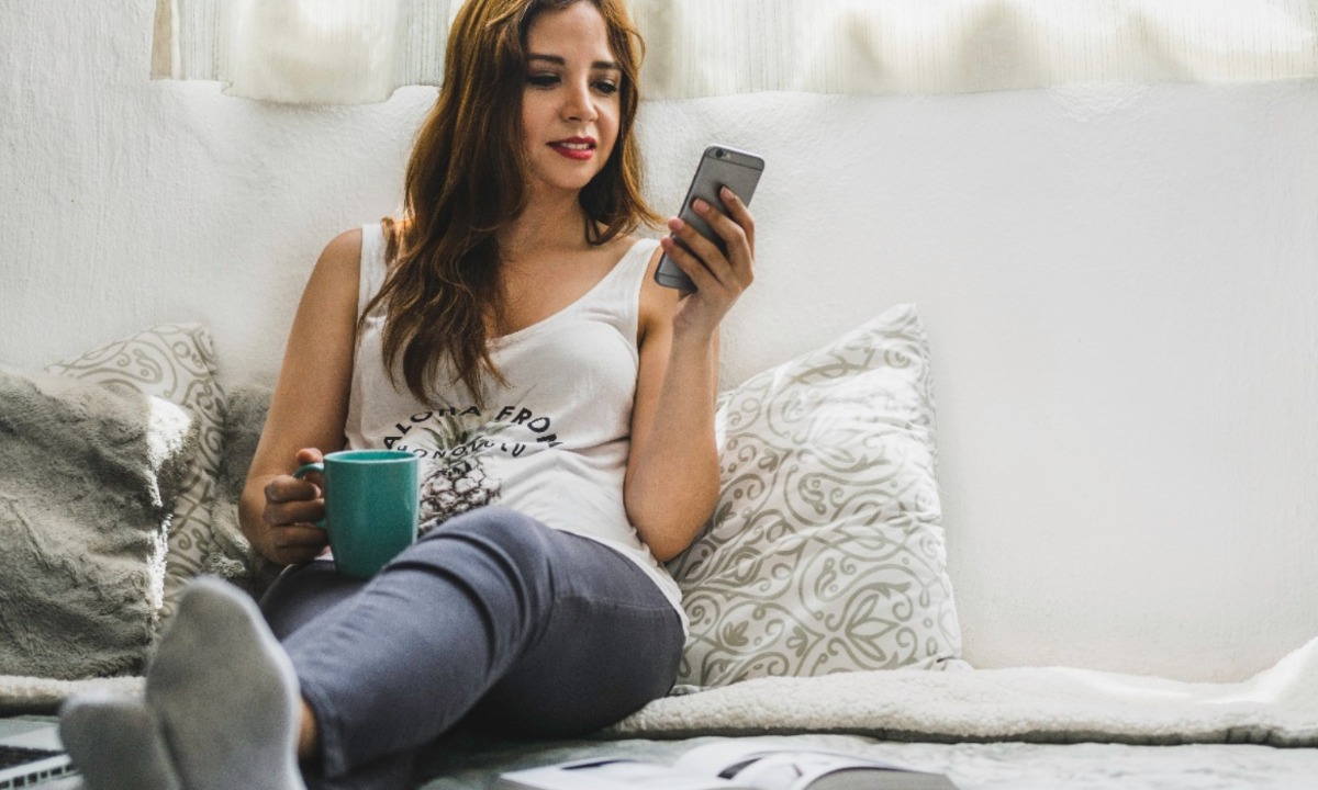 a woman sitting on a couch holding a phone and a cup of coffee