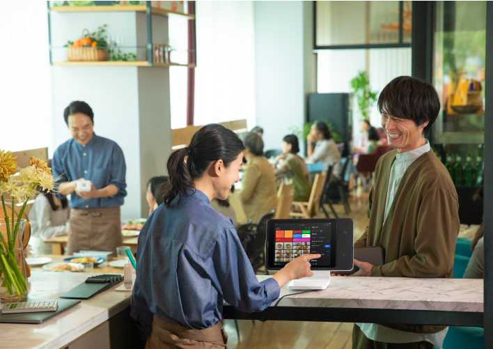 Two restaurant workers work on the register while handing a take-out bag to a delivery person to the right.