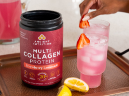 A container of Ancient Nutrition Multi Collagen Protein Strawberry Lemonade on a counter with a glass of it mixed in water with ice and a woman's hand.