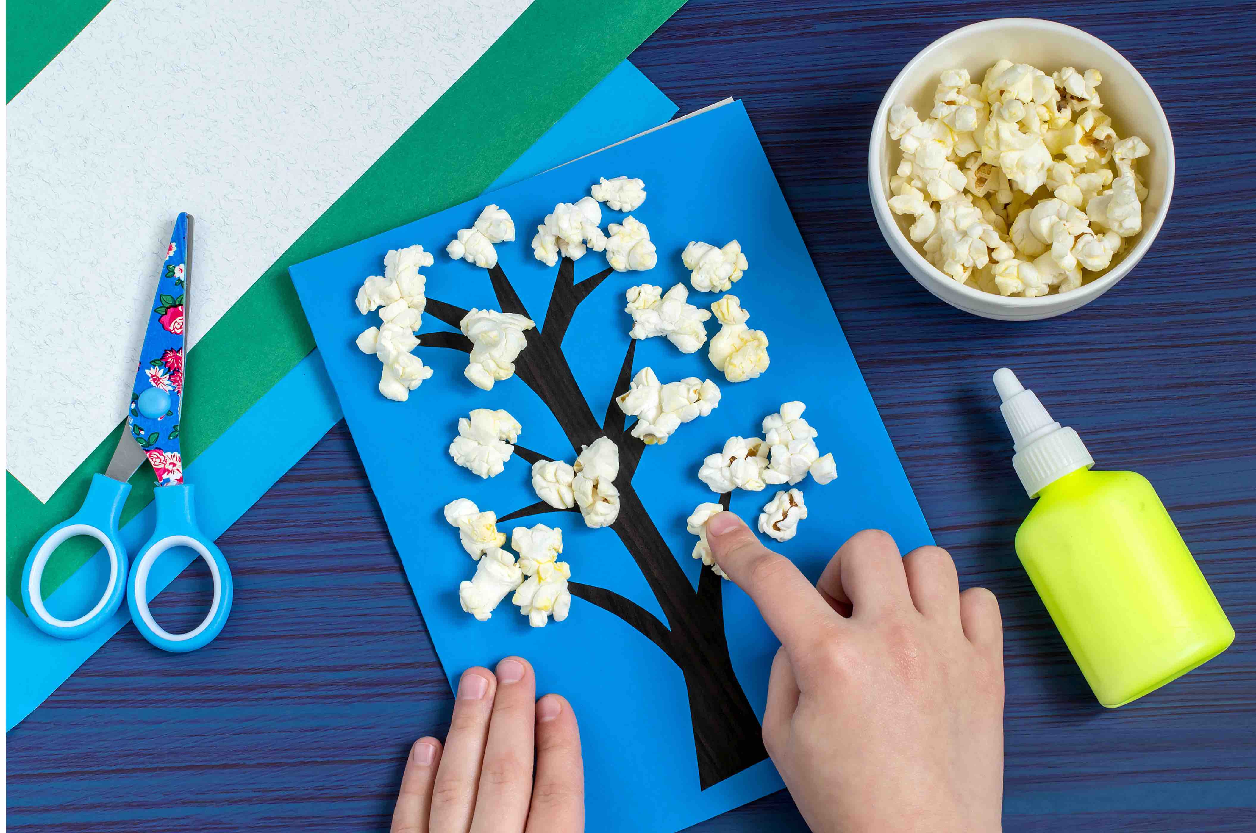 Making card by a child on spring theme. Blossoming tree on a blue background. Original children's art project. DIY concept. Step-by-step photo instruction. Step 5. Child glues flowers from popcorn By 13Smile - QF - 03