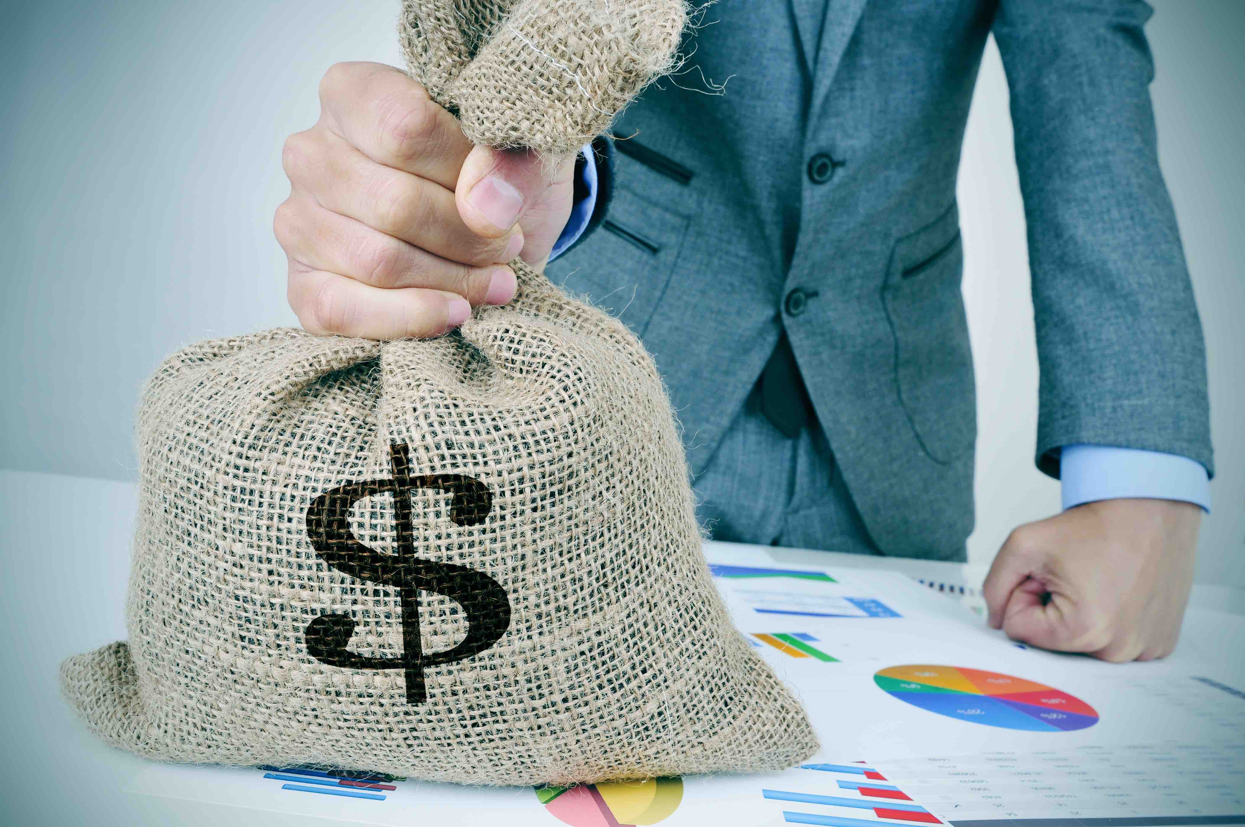 nito - money - share - closeup of a young caucasian man in a grey suit with a money bag on a desk full of different charts