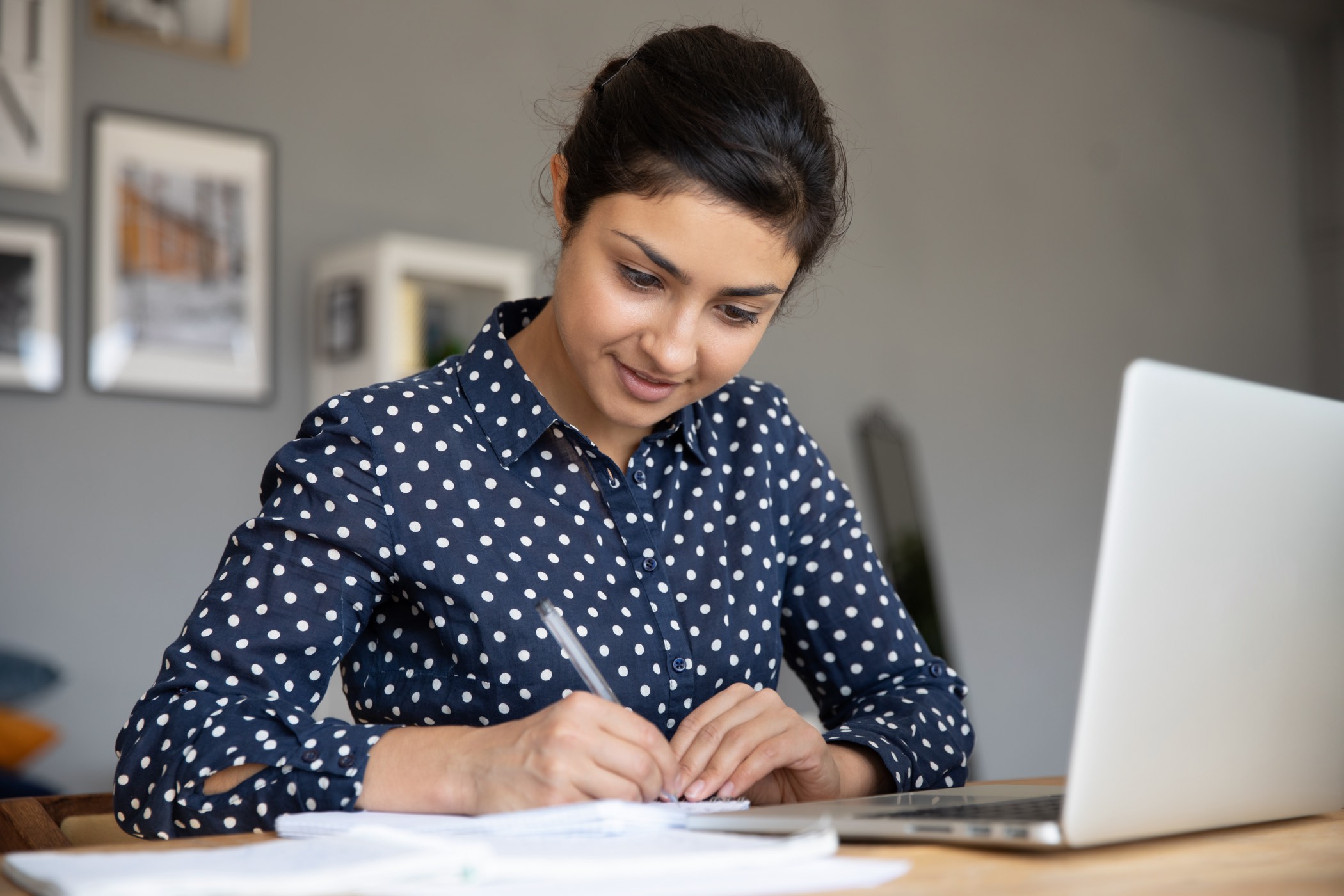 Confident indian businesswoman writing notes makes financial report sit in front of laptop at desk. Focused female working with paper documents, student studying on-line, creates research work concept - webinar - student -women with laptop computer