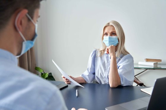 Female-hr-specialist-manager,-employer-wearing-face-mask-holding-cv-listening-male-candidate-at-job-interview-meeting.-Hiring-and-employment-during-covid-social-distance-safety-concept-By-insta photos