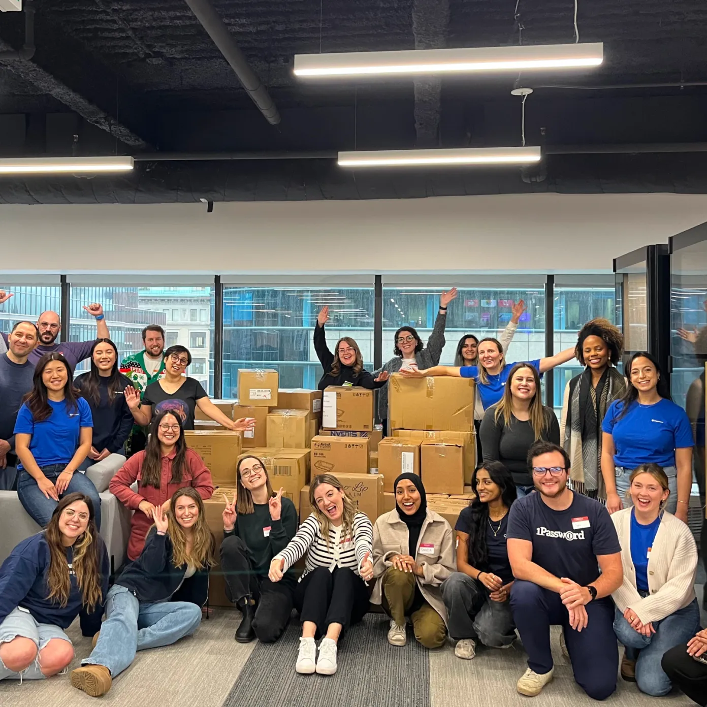 Group of 1Password employees gathered in an office in front of stacked donation boxes, smiling and waving.