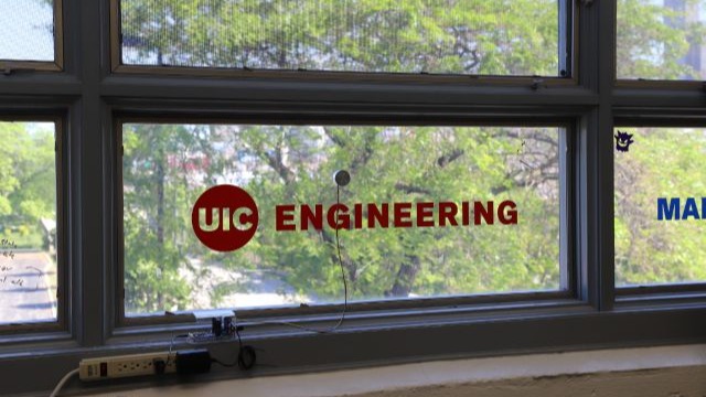 A window with the UIC Engineering school logo painted on the pane in front of a leafy green tree