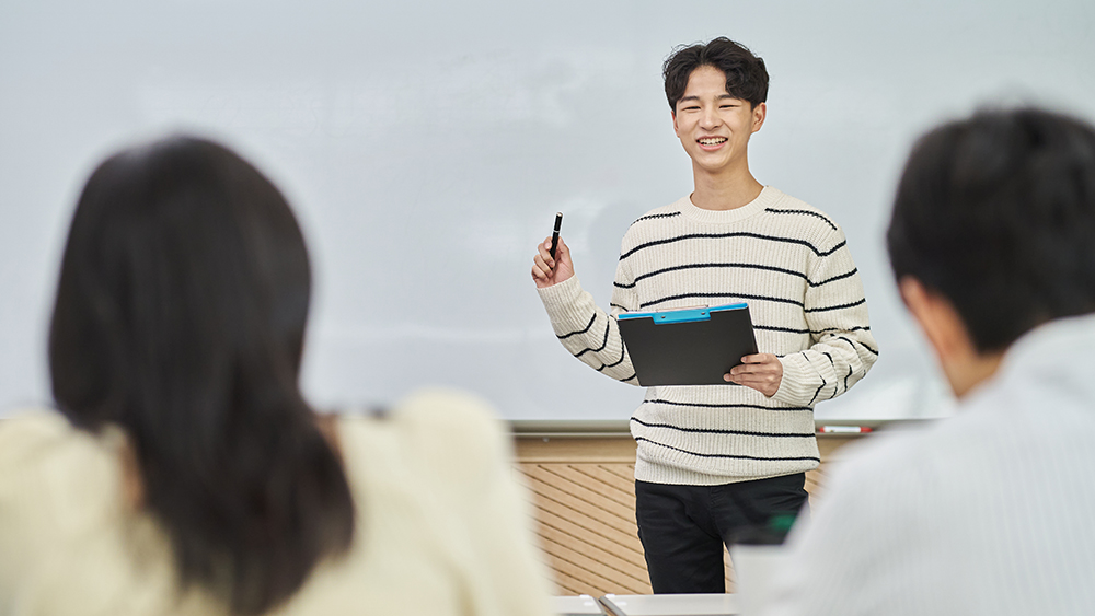 An Asian male international student stands in front of a whiteboard and presents his capstone project to his academic advisors.