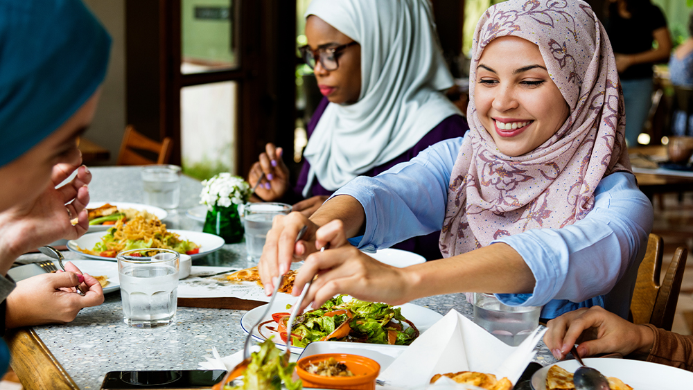 Three female international students wearing hijab sit at a table with salads and halal food at their US university dining hall.