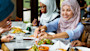 Three female international students wearing hijab sit at a table with salads and halal food at their US university dining hall.