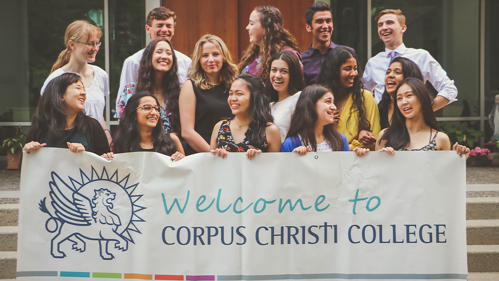 A group of international students stand on stairs on the UBC campus in Vancouver and hold up a banner saying Welcome to Corpus Christi College