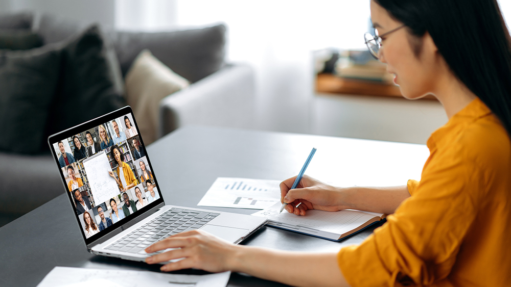 A female international student from Asia sits at her laptop and takes a US university class in a virtual classroom setting via a videoconference.
