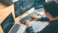 View from behind of a young man working at a desktop monitor and laptop, with both displaying computer code.