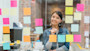 A female international student entrepreneur major stands in front of a glass wall covered in colorful sticky notes at her internship, working on answering what is a entrepreneur and listing the skills of entrepreneurs for her upcoming project.