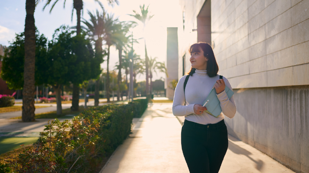 A female international student contemplates weather in America while walking under palm trees on her US university campus.