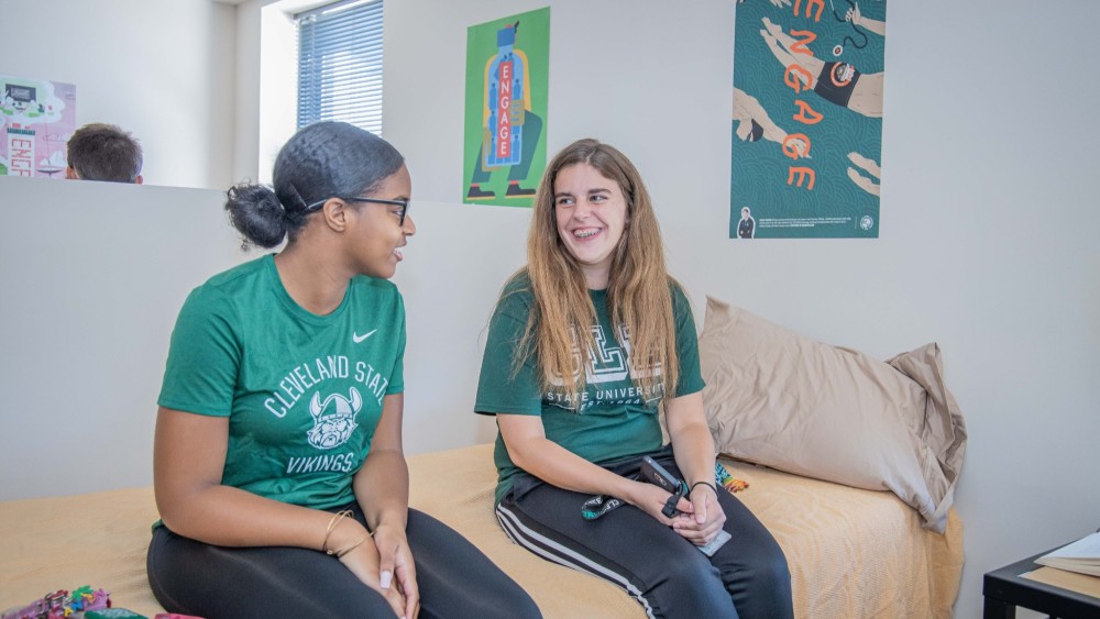 Two female international students laugh and talk while sitting side by side on a bed in a dorm at Cleveland State University