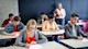 This photo shows university students taking the GMAT exam, with college students writing in GMAT test booklets with a female professor standing behind them.