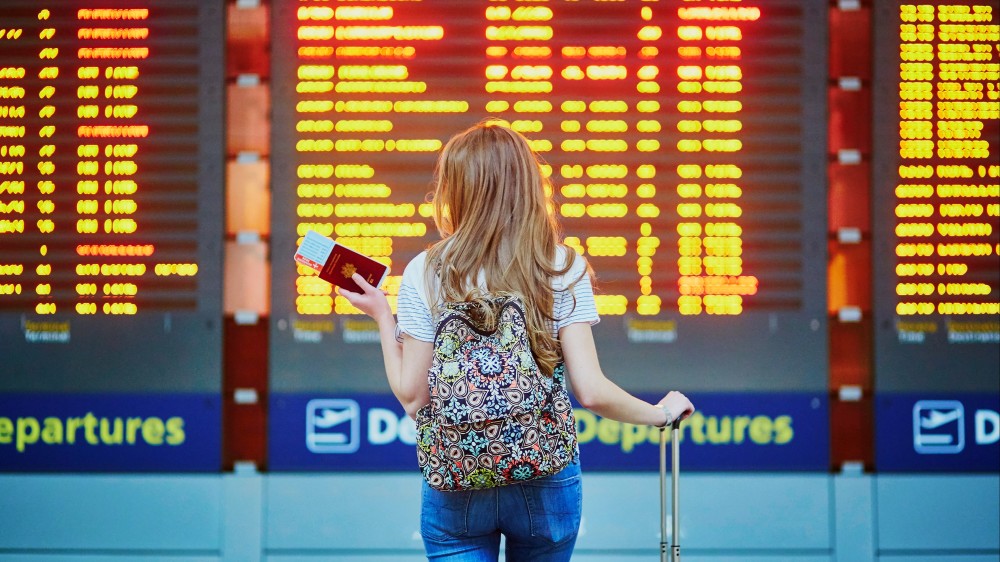 A female international student stands with a suitcase and her passport and j1 visa before a departures board at an airport