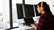 A female international student from Asia wearing headphones sits at her internship desk in front of windows showing a city skyline and codes on a laptop in front of two external monitors 