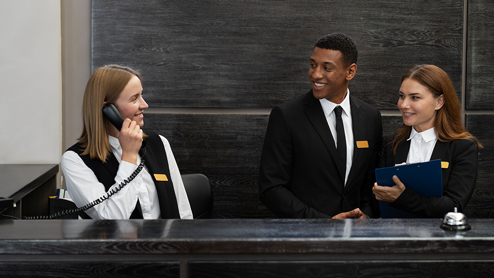 A hotel manager and two hotel management student intern employees work at the reception desk of a hotel. 