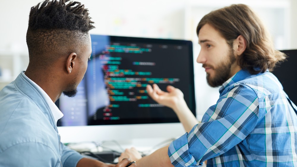 A hipster-looking guy gestures in front of a monitor displaying code during his technical interview