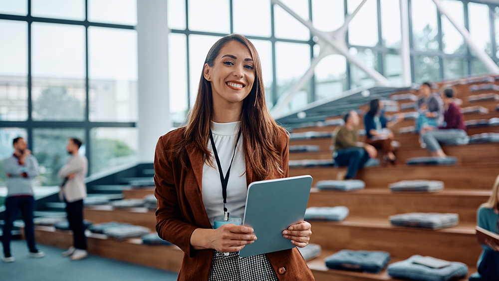 A female international student studying an event management course at her US university stands in a lecture hall before a big event, wearing an event lanyard and holding a tablet.