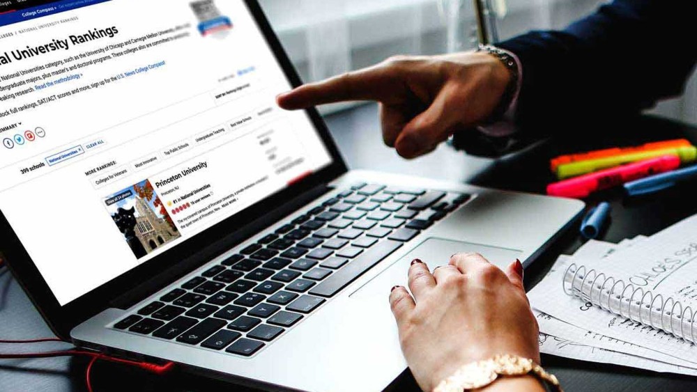 This photo shows an open laptop with a "us university rankings" article showing on the screen and a hand on the mousepad and a different hand pointing to the screen. The users are researching US top universities and US university ranking listings.  