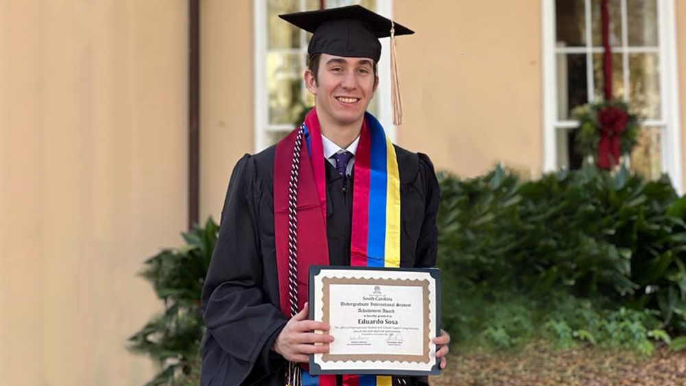 Eduardo, an international finance major from Chile, stands on the University of South Carolina campus in his graduation cap and gown holding the certificate for his International Student Achievement Award.