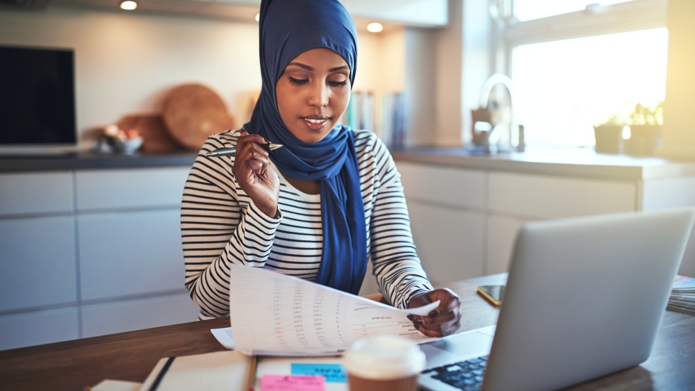 A female international student wearing a headscarf looks at a printed spreadsheet in front of a laptop at her kitchen table