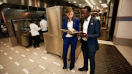 Two international students in uniform suits stand in a commercial kitchen and compare notes during their hospitality internship