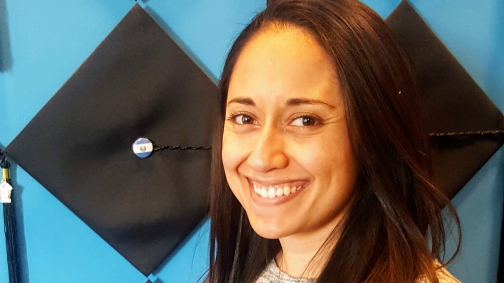 Amy Cruz, managing director at American Collegiate Los Angeles, stands in front of a wall of graduation caps and smiles for the camera