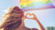 A female international student seen from behind holds her hands in a heart shape in front of a rainbow Pride LGBT flag