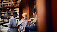 Three liberal arts education students have an animated discussion about the liberal arts, meaning the many different areas of study at their university, as well as their bachelor degree in liberal arts, while standing under the upper tier of books in a wood paneled library.