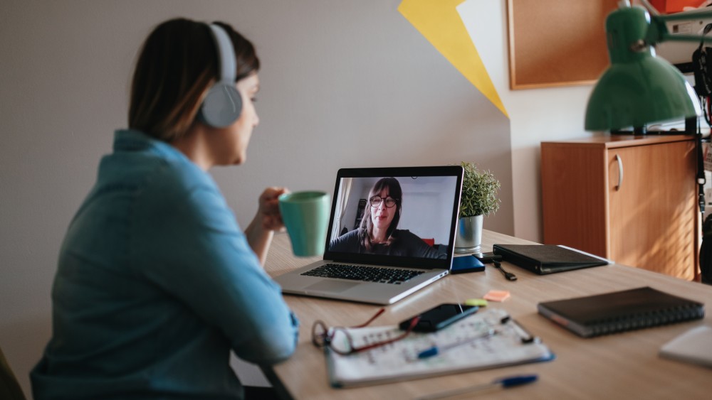 A female KU college student sits at a table in front of a laptop having a practice interview online with a female college counselor on the laptop screen