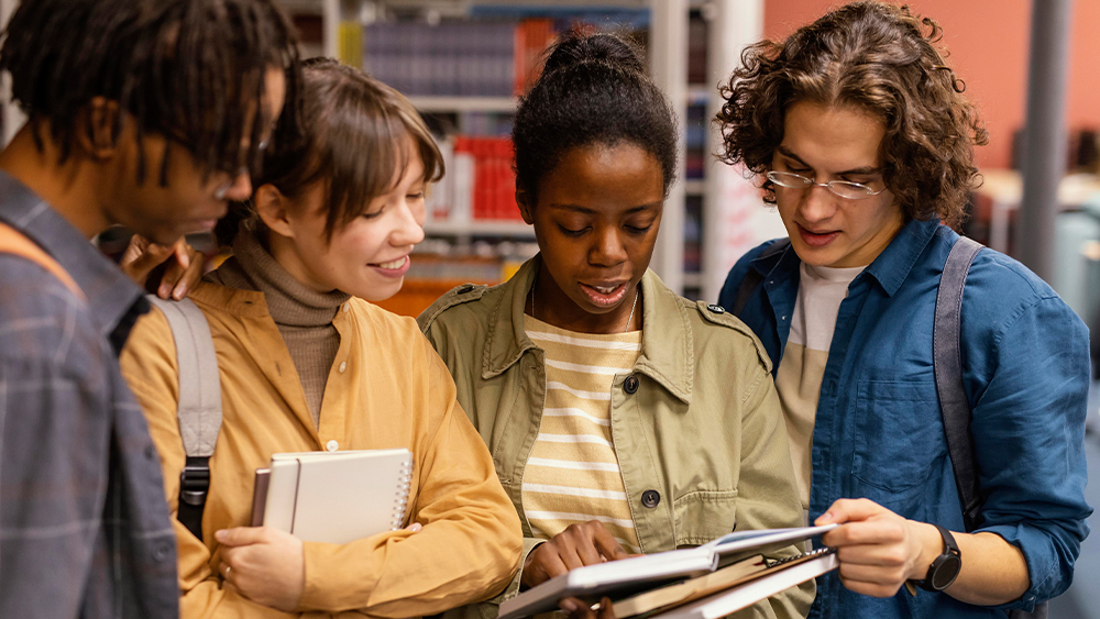 A group of four international students majoring in sociology at their US university stand in the library and consult the pages of a sociology textbook.