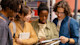 A group of four international students majoring in sociology at their US university stand in the library and consult the pages of a sociology textbook.